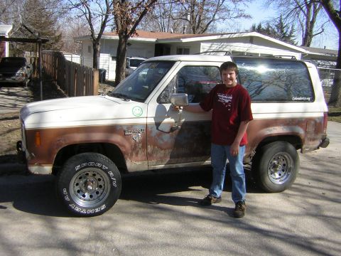 1986 Ford Bronco II - A boy stands next to a white and brown 1986 Ford Bronco II with visible rust and aftermarket wheels.