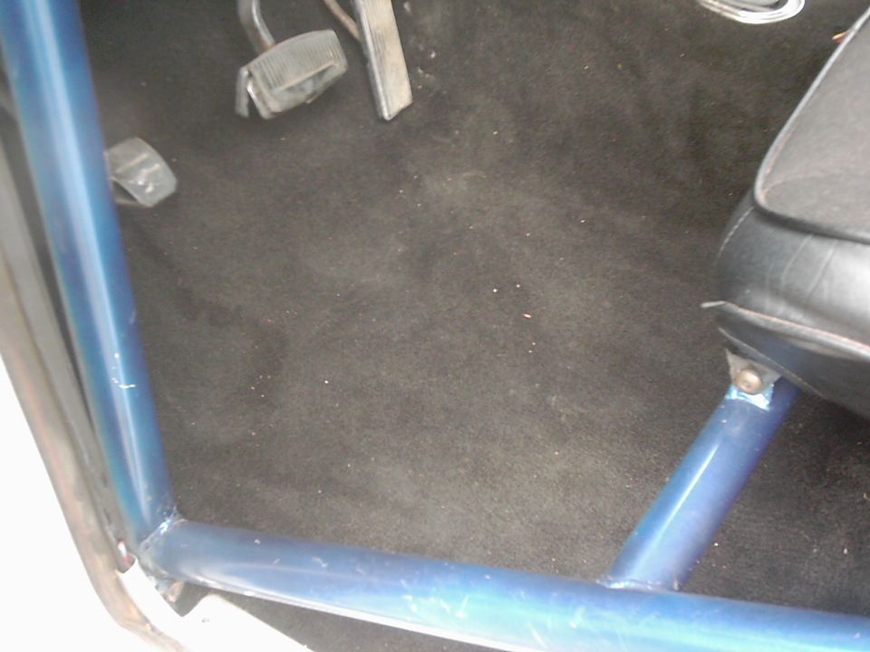 Vehicle interior showing black carpet, pedals, and a blue roll cage near the seat