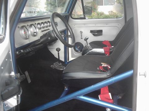 1978 Ford Bronco - Interior of a stripped truck cab shot through the open passenger door, showing black bucket seats with red racing harnesses, a blue tube ro…