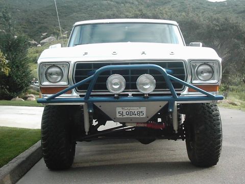 1978 Ford Bronco - Head-on front view of a lifted white late-1970s Ford 4x4 with FORD lettering across the hood, dual rectangular headlights, and a blue tubul…