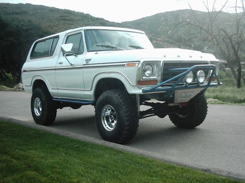 1978 Ford Bronco - White lifted full-size Ford Bronco photographed from the front three-quarter angle on a paved driveway with green hills behind it. It wears…