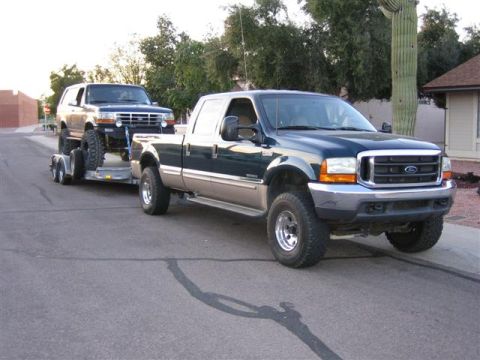 1999 Ford F-250 - A 1999 Ford F-250 Super Duty tows a lifted Ford Bronco on a trailer down a residential street.