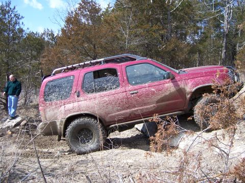 1999 Jeep TJ - A red SUV tackles a challenging off-road course with a person observing nearby.