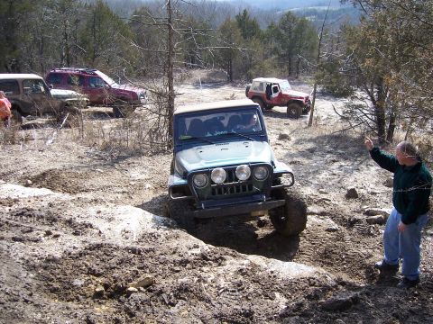 1999 Jeep TJ - Multiple Jeeps tackle an off-road trail with muddy terrain and sparse trees.