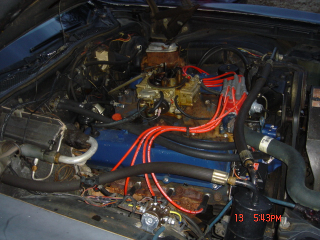 Blue V8 engine bay with gold four-barrel carburetor and red plug wires