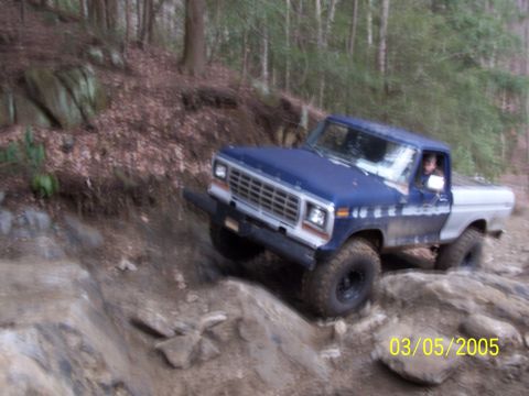 1979 Ford F-150 - A blue and silver 1979 Ford F-150 navigates a challenging rocky terrain on a wooded trail.