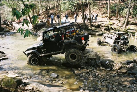 1979 Ford F-150 - Off-road vehicles are shown traversing a shallow, rocky stream, with onlookers observing from the wooded bank.