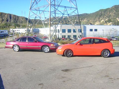 1992 Ford Crown Victoria - A red early-1990s Ford Crown Victoria sedan and a bright orange first-generation Ford Focus five-door hatchback parked nose to nose in a pa…