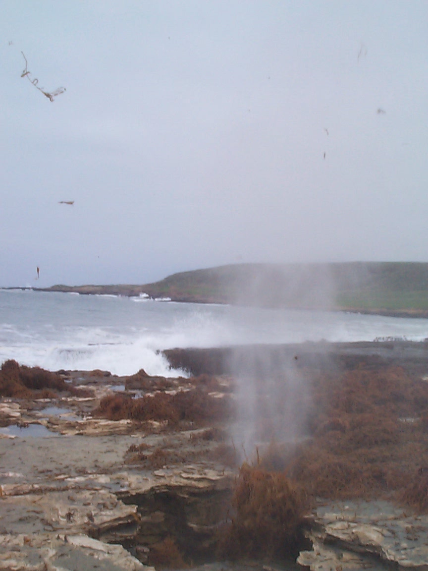 Rocky shoreline with surf spray and sea mist drifting over a flat gray coastal bay