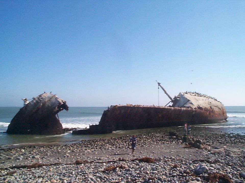 Large rusted shipwreck broken in two halves resting on a pebble beach with birds on top