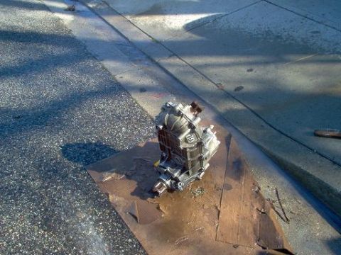 1988 Ford Bronco - A transfer case sits on a piece of cardboard on the asphalt, with some text visible on its housing.