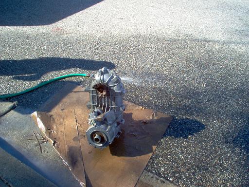 Aluminum transfer case housing being washed with a garden hose on plywood in a driveway