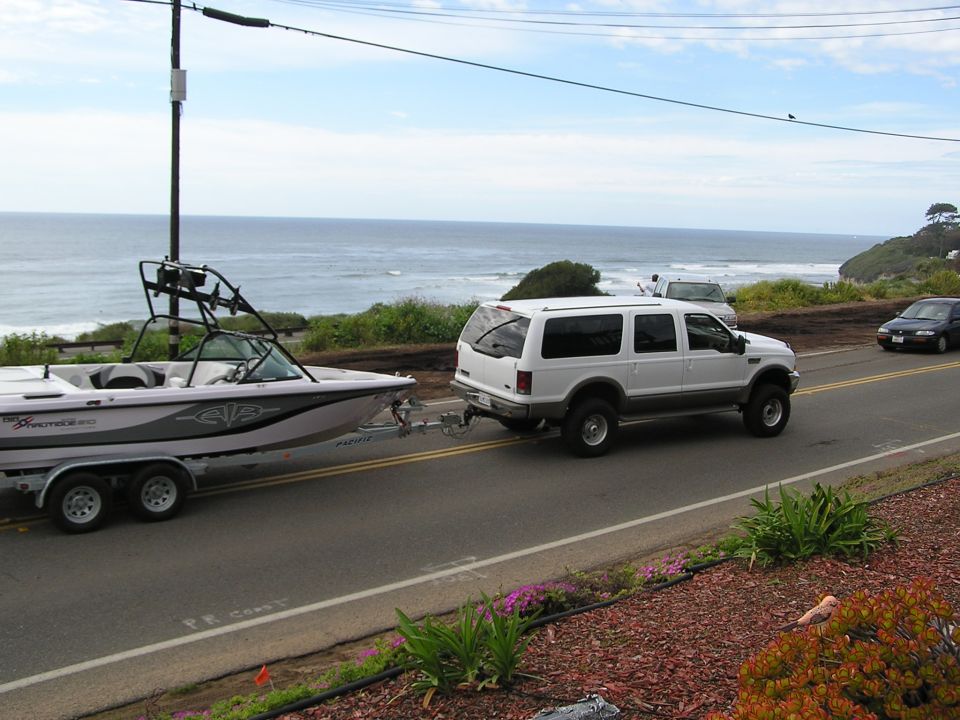 White Ford Excursion SUV towing a boat on a trailer along a coastal road by the ocean.