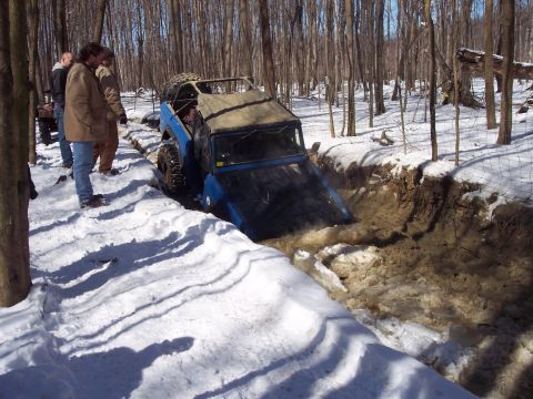 1974 Ford Bronco - A blue off-road vehicle navigates a challenging muddy ditch surrounded by snow and trees.