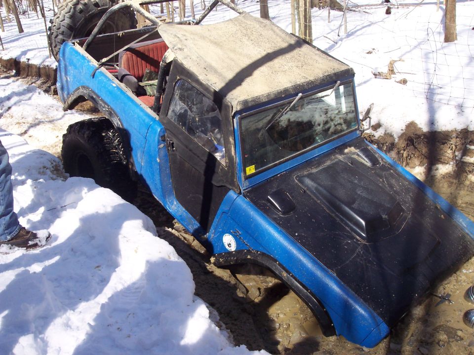 Blue off-road vehicle with a black hood stuck in a muddy ditch with snow on the ground