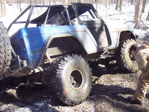 1974 Ford Bronco - A modified 1974 Ford Bronco with a roll cage and oversized tires navigates a muddy, wooded area.