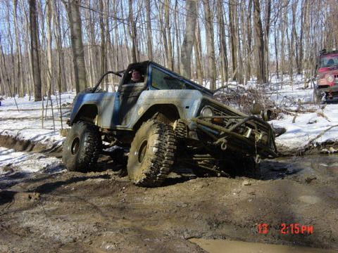 1974 Ford Bronco - A heavily modified off-road vehicle navigates a muddy trail in a wooded area with snow on the ground.