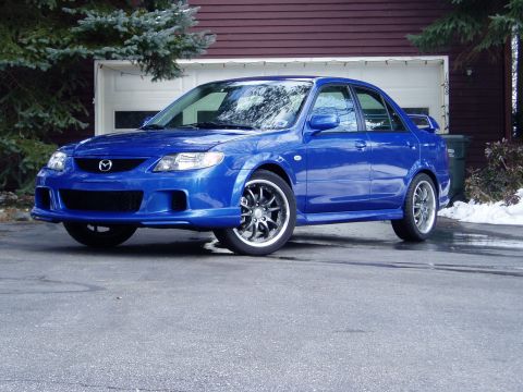 2003 Mazda Protege - This blue 2003 Mazda Protege sedan features an aftermarket body kit and custom wheels, seen here parked in front of a garage.