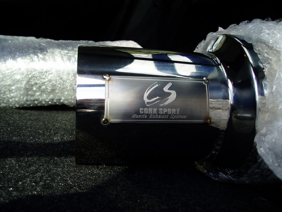 Polished CorkSport Mazda exhaust tip with engraved badge, wrapped in bubble wrap on dark carpet
