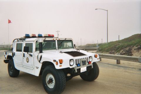 1990 Ford Bronco - white Hummer H1 fitted with a roof light bar sits on pavement beside a guardrail in coastal fog.