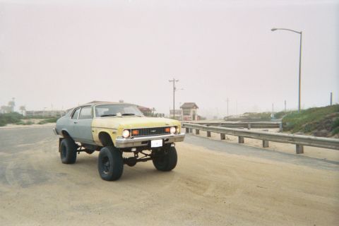 1990 Ford Bronco - lifted blue-and-yellow Chevy Nova coupe sits high on solid axles and big tires on a misty paved beach road.