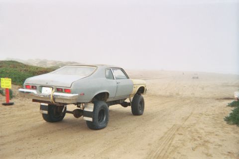 1990 Ford Bronco - Rear three-quarter view of a lifted gray-and-yellow Nova coupe raised on tall tires on sandy dune ground in fog.