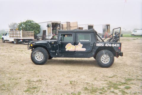 1990 Ford Bronco - black soft-top Hummer with business lettering and phone number parked in a foggy dirt lot near a flatbed truck.