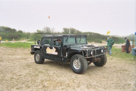 1990 Ford Bronco - black soft-top Hummer H1 with off-road tires sits on a fog-shrouded beach as people stand near a table in the background.