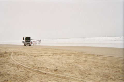 1990 Ford Bronco - lone vehicle tows gear along a wide expanse of tire-tracked beach beside the breaking surf under hazy skies.