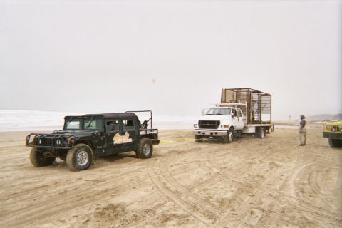 1990 Ford Bronco - black Hummer and a white flatbed truck with a cage sit on a tire-marked beach as a person stands nearby in the fog.