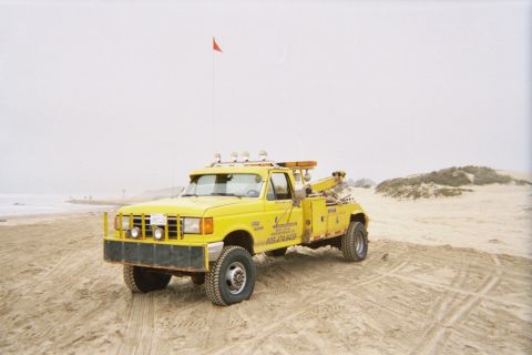 1990 Ford Bronco - yellow lifted 4x4 dually tow truck with a roof light bar sits on a sand dune beach under a hazy sky with a red flag.