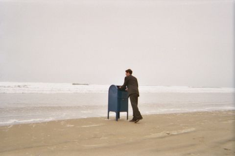 1990 Ford Bronco - barefoot man in a dark suit leans over a blue mailbox at the surf line on a wide empty beach.