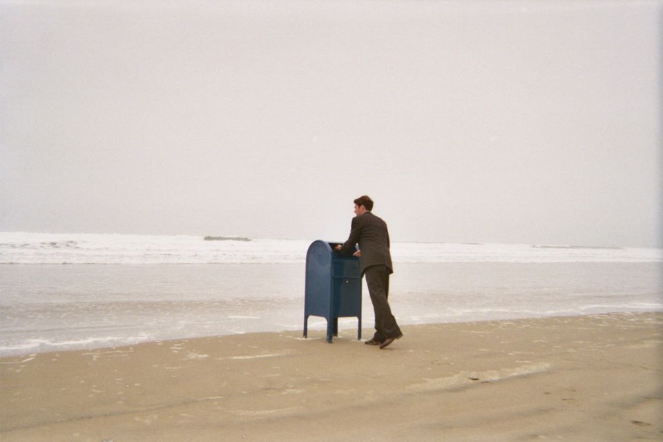 Man in a suit using a blue mailbox on the wet sand at the water's edge