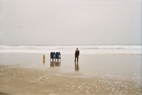 1990 Ford Bronco - man in a dark suit stands at the water's edge beside two blue mailboxes and a yellow hydrant on a misty beach.