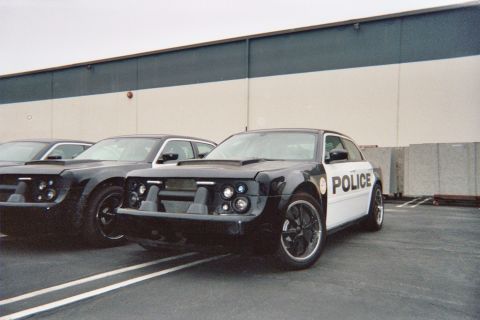 1990 Ford Bronco - black-and-white POLICE-liveried custom sedan with a blacked-out front sits in a lot beside a similar black car.