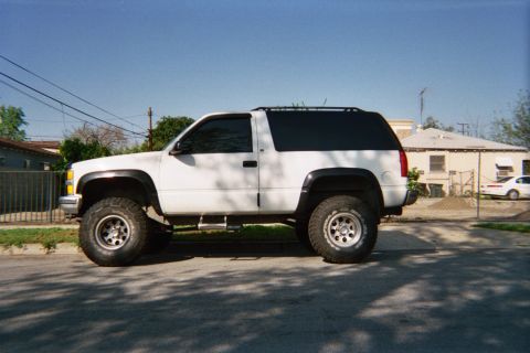 1990 Ford Bronco - white lifted two-door Chevrolet Tahoe with fender flares and off-road tires sits curbside on a sunny residential street.