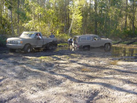 1989 Ford Bronco - Two SUVs are stuck in a muddy, wooded area, with one vehicle attempting to tow the other out.