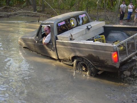 1989 Ford Bronco - A pickup truck is stuck in a muddy body of water with people watching in the background.
