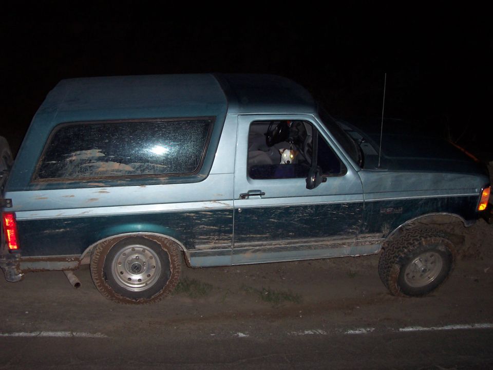 Side view of a two-tone green and blue Ford Bronco with a camper shell, covered in mud