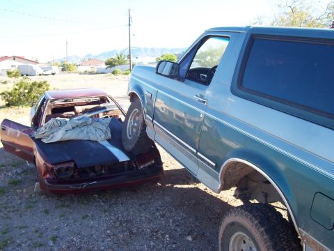 1996 Ford Bronco - A blue SUV is parked on top of a wrecked red car in a dry, outdoor area.