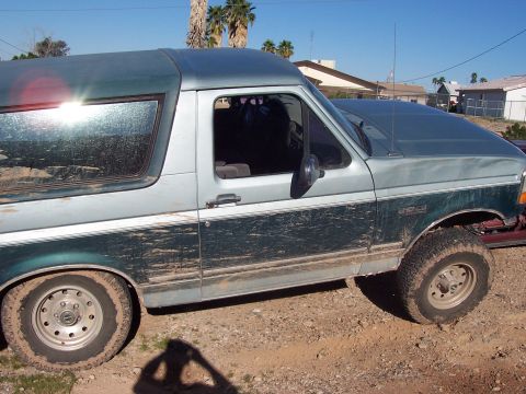 1996 Ford Bronco - A muddy two-tone green and silver Ford Bronco is parked on dirt ground under a clear sky.