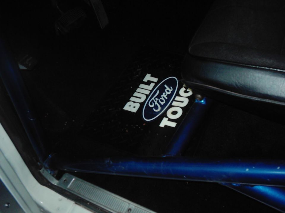 Interior shot of a vehicle showing a black floor mat with "BUILT Ford TOUGH" text and logo. Blue roll cage visible