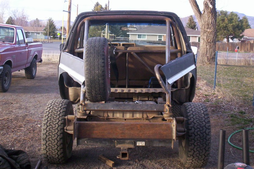 1983 Ford Bronco rear view with custom roll cage, spare tire mounted on back, and a red Ford pickup truck in the background