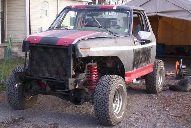 Modified pickup truck with red and black stripes, lifted suspension, and large tires. Front end is partially disassembled