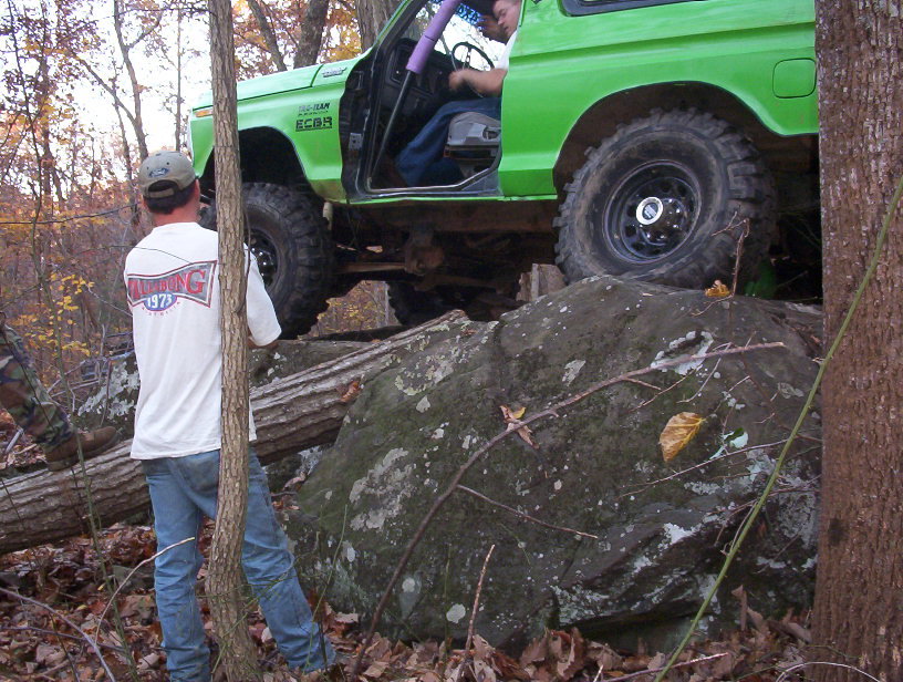 Green door-off Bronco perched on a large boulder in fall woods with spotter watching