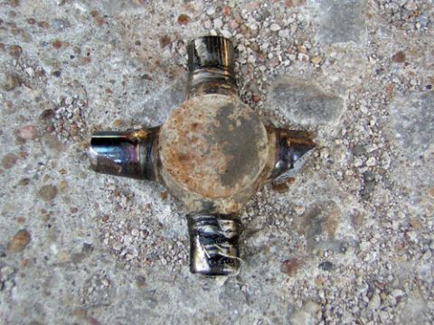 1995 Ford Bronco - A worn universal joint (U-joint) cross with four bearing caps, showing rust on the central body and shiny metal caps, laid on a concrete su…