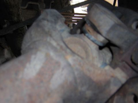 1995 Ford Bronco - A tight, dimly lit close-up of a rusty cast undercarriage component, likely a front axle knuckle or differential area with blue paint trace…