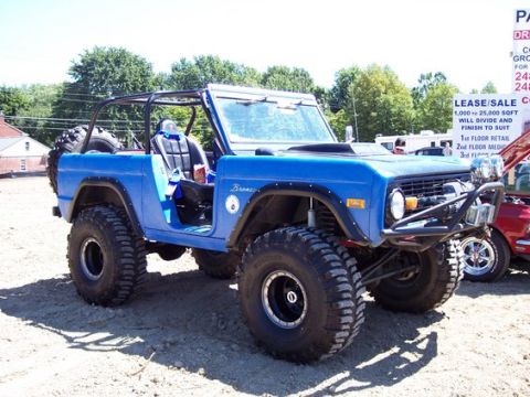 1990 Ford Bronco - blue lifted early Ford Bronco with an open top, roll cage and oversized tires sits on gravel at an outdoor car show.