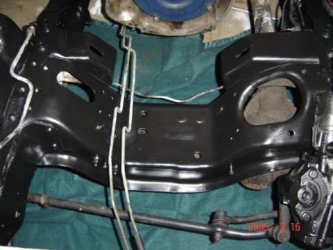 1990 Ford Bronco - Overhead shot of the freshly painted gloss-black engine crossmember and frame rails in an empty engine bay, with white brake hard lines rou…