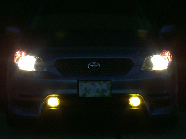Front of silver Toyota at night with headlights and yellow fog lights on
